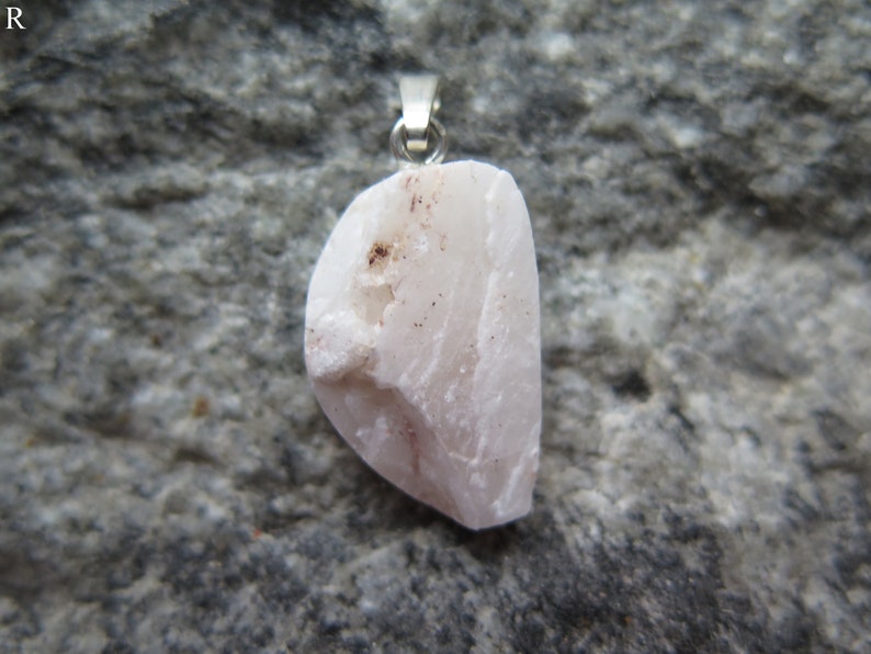 May include: A pale pink, irregularly shaped stone pendant with a silver bail. The stone has a rough, natural texture and subtle red veins.
