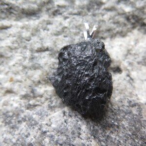 May include: A black, irregularly shaped pendant with a textured surface. The pendant has a silver-colored bail at the top for a necklace. The pendant is set against a gray, textured background.