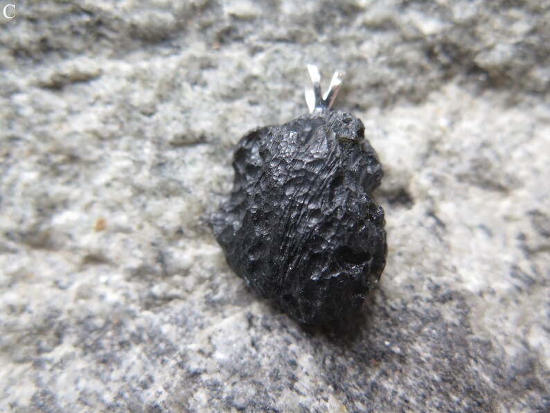 May include: A black, irregularly shaped pendant with a textured surface. The pendant has a silver-colored bail at the top for a necklace. The pendant is set against a gray, textured background.