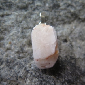 May include: A pale pink, irregularly shaped stone pendant with a silver bail. The stone has a few reddish-brown inclusions.