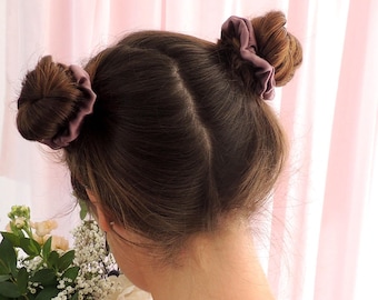May include: A person's hair styled in two buns, each secured with a mauve scrunchie. The hair is brown, and the person is facing away from the camera. A bouquet of white and green flowers is visible in the foreground, with a pink curtain in the background.