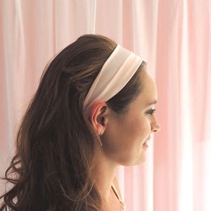 Beth, a white woman in her mid twenties, is facing to the side towards a pink fabric background with her brunette hair held back by a pale pink silk headband as a styling example