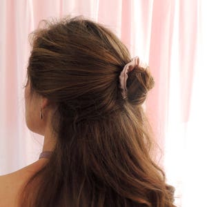 Puede incluir: Cabello castaño peinado en un peinado recogido a medias. Una goma de pelo rosa claro asegura la sección superior. El cabello es ondulado y cae sobre los hombros. Se ve un pendiente delicado.