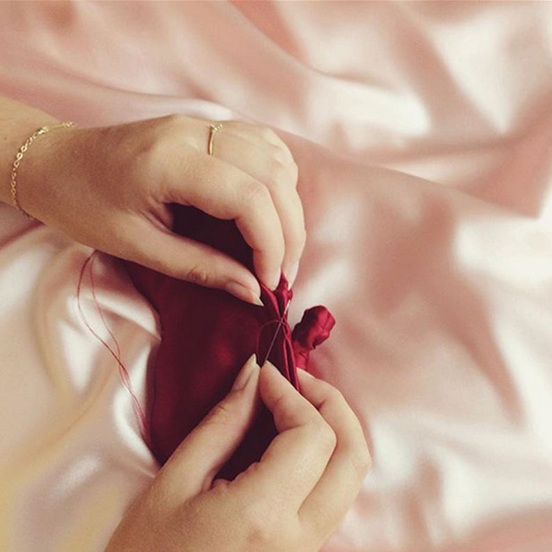 A deep red real silk eye mask with two hands sewing a seam closed against a pale pink silk background