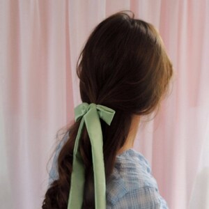 Beth is facing away from the camera towards a pale pink fabric backdrop. Her brunette hair is tied half up with a pale mint green silk ribbon