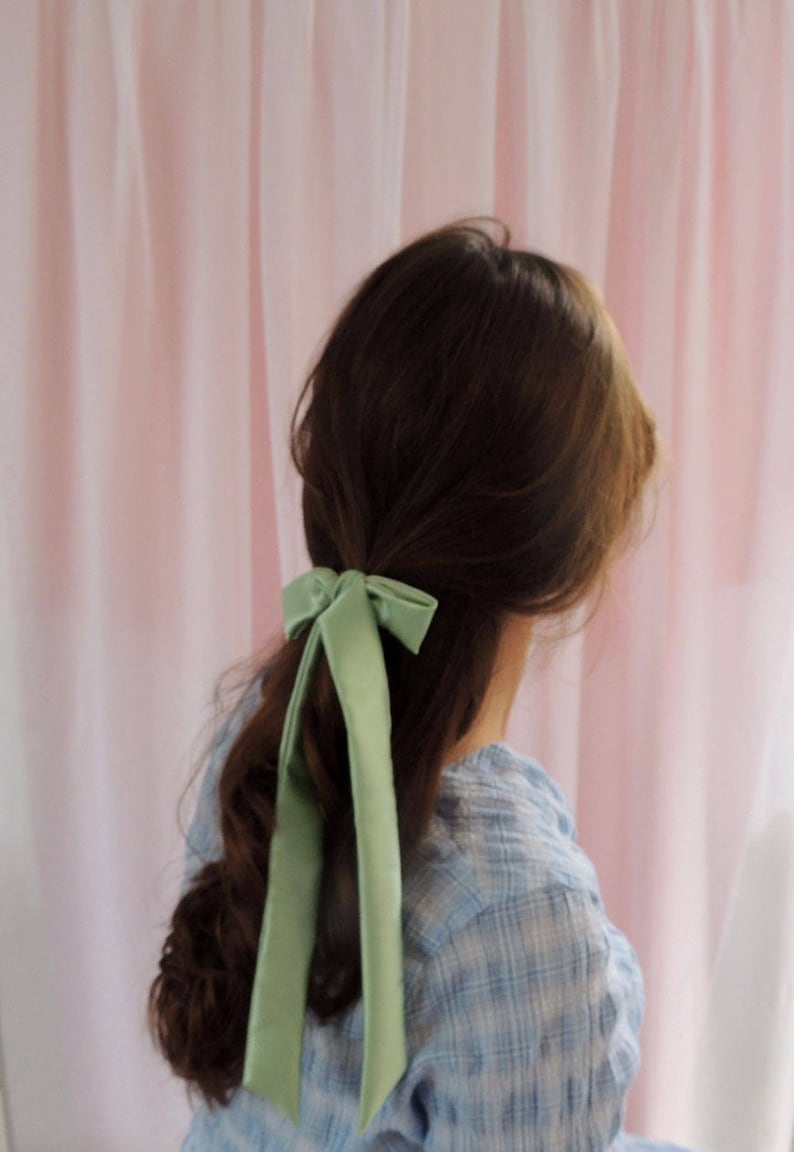 Beth is facing away from the camera towards a pale pink fabric backdrop. Her brunette hair is tied half up with a pale mint green silk ribbon