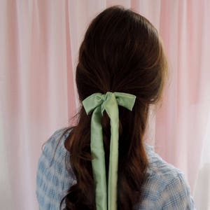 Beth is facing away from the camera towards a pale pink fabric backdrop. Her brunette hair is tied half up with a pale mint green silk ribbon