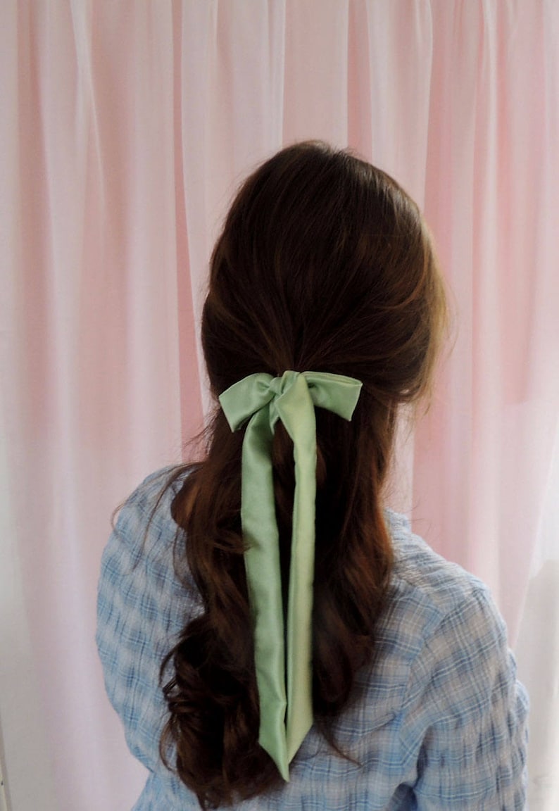 Beth is facing away from the camera towards a pale pink fabric backdrop. Her brunette hair is tied half up with a pale mint green silk ribbon
