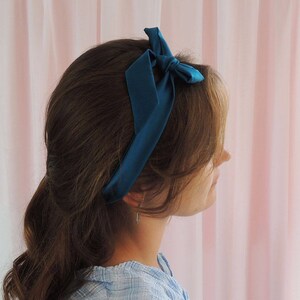 Beth, a white woman, is facing away from the camera towards a pale pink fabric backdrop. Her brunette hair has a deep teal silk ribbon tied around in a headband style