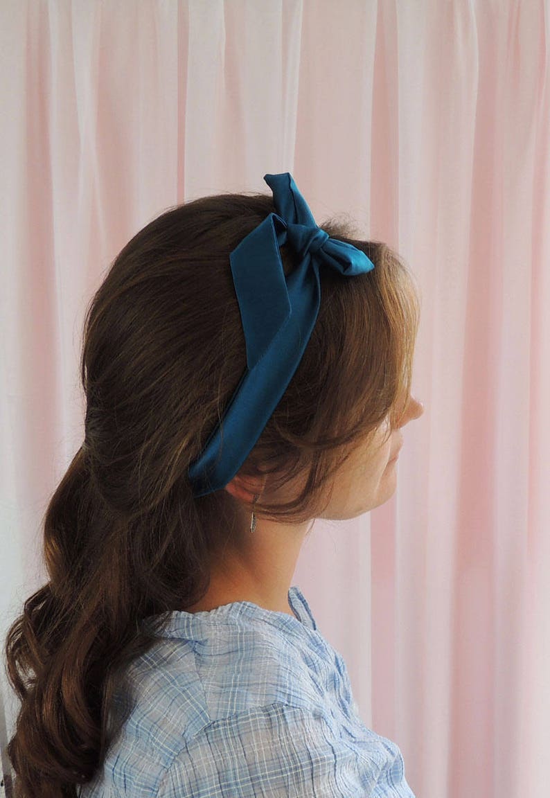 Beth, a white woman, is facing away from the camera towards a pale pink fabric backdrop. Her brunette hair has a deep teal silk ribbon tied around in a headband style