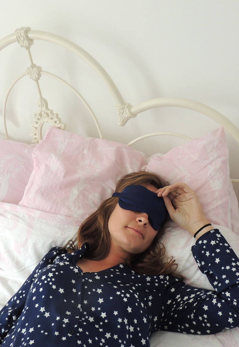 Beth, a white woman in her mid twenties with long brunette hair, wears a navy blue silk eye mask wearing navy star print pyjamas on a white metal frame bed with pink bedding