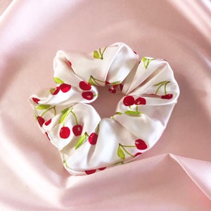 Könnte beinhalten: Ein weißer Satin-Haargummi mit Kirschendruck. Der Haargummi ist zusammengezogen und zu einem Knoten gebunden.