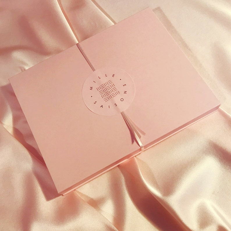 A light pink cardboard box sealed with ribbon and a Mille Saisons logo sticker on a pale pink silk background
