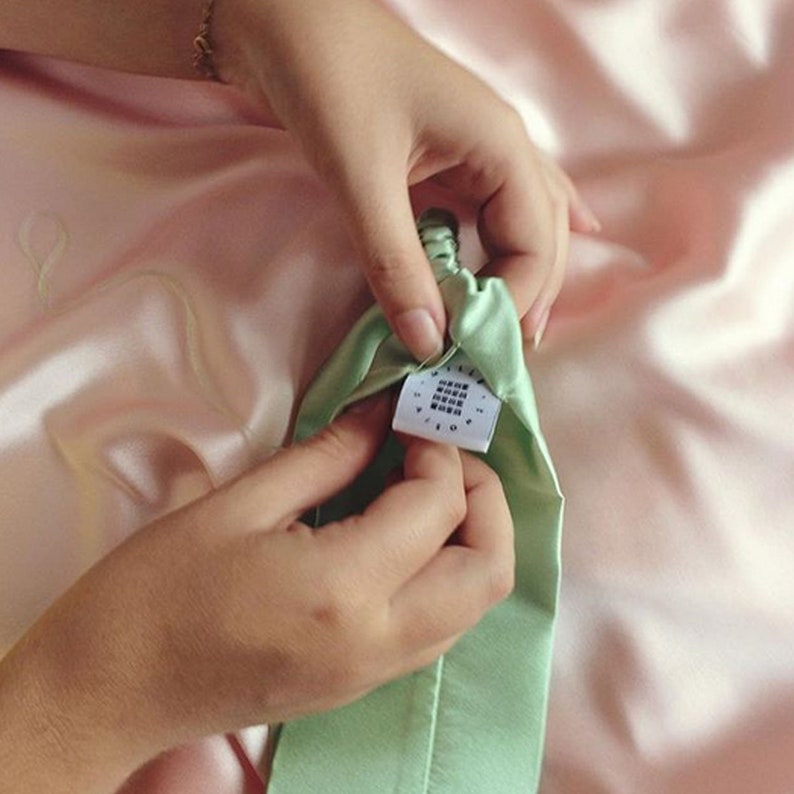 A close up of a pale green silk headband with elasticated silk covered strap and Mille Saisons logo tag being hand sewn into the side against a pale pink silk background