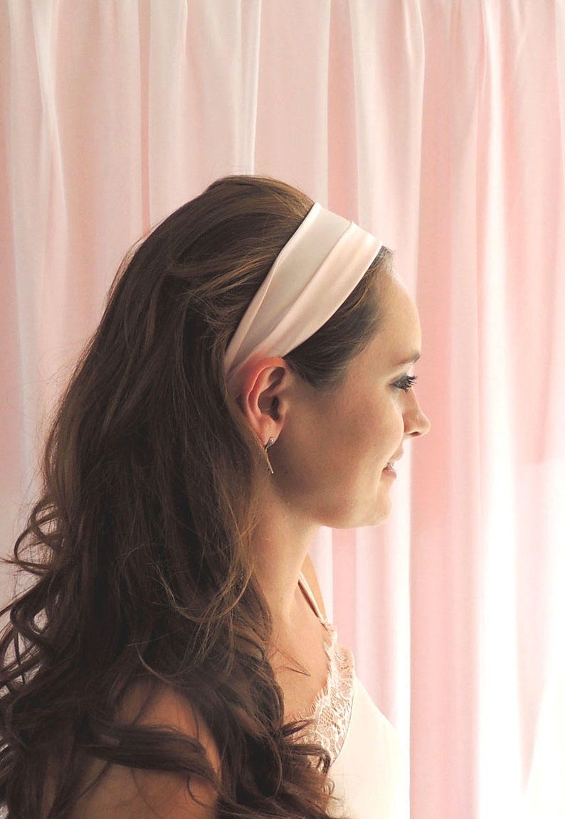 Beth, a white woman in her mid twenties, is facing to the side towards a pink fabric background with her brunette hair held back by a pale pink silk headband as a styling example