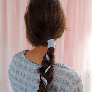 Beth, a white woman, is facing away from the camera towards a pale pink fabric backdrop. Her brunette hair is in a loose plait woven with a pale blue silk ribbon