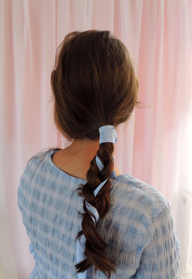 Beth, a white woman, is facing away from the camera towards a pale pink fabric backdrop. Her brunette hair is in a loose plait woven with a pale blue silk ribbon