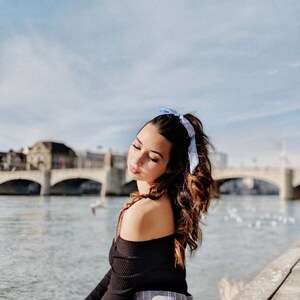 Jordan, a white woman,  is sat by a river. Her brunette hair is tied up with a pale blue silk ribbon
