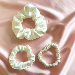 Two silk scrunchies in ivory and a narrower silk hair elastic showing the difference in size between a classic size, hair elastic and mini size