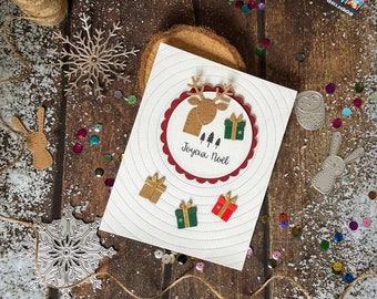 May include: A handmade Christmas card with a reindeer and gift design. The card features the text "Joyeux Noel" and is decorated with gold, green, and red gift embellishments. The card is on a wooden surface with decorative elements.