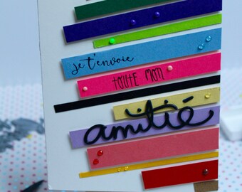 May include: A handmade greeting card on a small wooden easel. The card features colorful horizontal stripes in orange, green, blue, and pink, with the French text "je t'envoie toute mon amitié" in black script. Decorative embellishments include small colored dots.