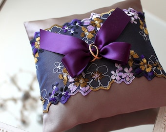 May include: A brown satin pillow with a purple ribbon and floral lace trim. Two gold rings are tied to the ribbon.