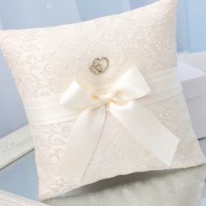 May include: Ivory lace ring pillow with a satin ribbon and bow. A silver charm with two intertwined hearts is centered on the pillow. The pillow is square and sits on a reflective surface, with a white box in the background.