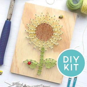 May include: A DIY kit featuring a sunflower string art design on a wooden board. The sunflower is crafted with yellow and brown thread, with green leaves and stem. The kit includes a hammer, nails, and the text "DIY KIT" in a teal circle.