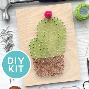 May include: A DIY kit featuring a cactus string art design. The cactus is crafted with green thread and silver pins, set against a wooden board. The pot is made with brown thread, and a pink pom-pom tops the cactus. The words "DIY KIT" are in a blue circle.
