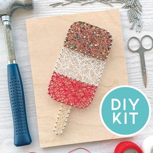 May include: A DIY string art kit depicting a popsicle design. The popsicle is made with red, white, and brown string on a wooden board. A hammer, scissors, and nails are visible, with the text "DIY KIT" in a teal circle.