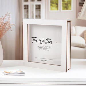 Könnte beinhalten: Eine weiße Schattenbox mit einer transparenten Frontplatte. Der Text "The Watters" ist in einer Schreibschrift geschrieben, mit einem Zweigdesign und dem Datum "EST. 2025" darunter. Die Box hat eine braune Umrandung und steht auf einer weißen Oberfläche.