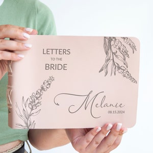 Pode incluir: Um di&aacute;rio de couro rosa com o texto "Cartas para a noiva" e o nome "Melanie" com a data "08.15.2024" na capa. O di&aacute;rio tem um design floral com um delicado desenho de flores.