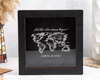 May include: A black framed world map with the text "let the adventures begin" and the names "Martha & David". The map is white and the frame is square. The background is black.