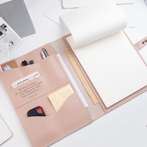 May include: A light pink leather-like portfolio organizer featuring multiple interior pockets, a notepad, and a snap closure. The left panel displays the printed text: Be strong and courageous. Do not be afraid; do not be discouraged, for the Lord your God will be with you wherever you go. JOSHUA 1:9.