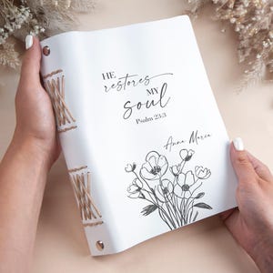 May include: White journal with the text "He restores my soul" and "Psalm 23:3". The journal has a floral illustration and is bound with tan twine. The journal is held open by a person.