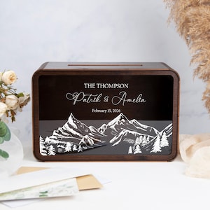 May include: A dark brown wooden box with a slot on top, decorated with a mountain and forest scene. The front panel displays the names "Patrik & Amelia" and the date "February 15, 2026". This box is likely for a wedding or special event.