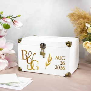 May include: White wooden card box with gold-colored corner protectors and accents. The box features gold lettering with the initials "B&G" and the date "AUG 26 2026". A small lock and key are attached to the front.