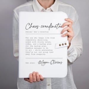 Könnte beinhalten: Ein weißer, rechteckiger Planer mit Druckknopfverschluss, bedruckt mit dem Text Chaos coordinator, einer phonetischen Aussprachehilfe und einer humorvollen Definition über die Lebensplanung, abgeschlossen mit dem Namen Megan Oliviers in Schreibschrift.