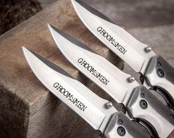 May include: Three silver pocket knives with wooden handles. Each knife has the word "Groomsmen" engraved on the blade.