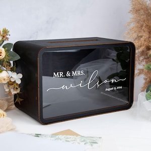 Könnte beinhalten: Eine schwarze Holzkartenbox mit einer transparenten Frontplatte. Die Box hat abgerundete Ecken und einen Schlitz oben. Weißer Text auf der Vorderseite lautet "MR. & MRS. wilson August 15, 2024".