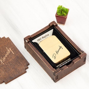 May include: A gold-colored lighter with the inscription "D. Kennedy" in a wooden box with a black velvet lining. The box is engraved with the text "D. Kennedy" and the words "1941 Replica" are visible on a piece of paper inside the box.