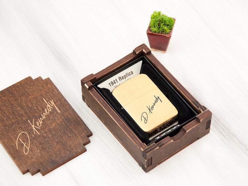 May include: A gold-colored lighter with the inscription "D. Kennedy" in a wooden box with a black velvet lining. The box is engraved with the text "D. Kennedy" and the words "1941 Replica" are visible on a piece of paper inside the box.