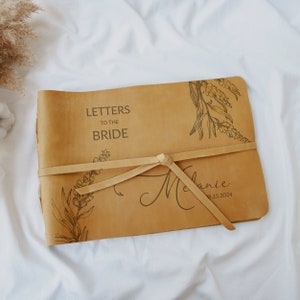Pode incluir: Um di&aacute;rio encadernado em couro castanho com um design floral e o texto "Letters to the Bride" e "Melanie 08.15.2024" gravado na capa.