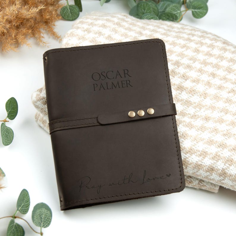 K&ouml;nnte beinhalten: Ein braunes Lederjournal mit dem Namen "Oscar Palmer" auf dem vorderen Umschlag gepr&auml;gt. Das Journal hat einen Riemenverschluss mit goldfarbenen Nieten und die Worte "Pray with love" sind unten eingraviert.