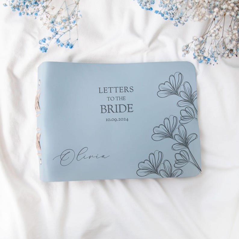 May include: A light blue leather-bound journal with the text "Letters to the Bride" and the date "10.09.2024" printed on the front cover. The name "Olivia" is written in cursive below the date. The cover features a delicate floral design in gray.