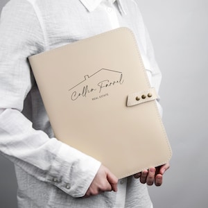 May include: A beige document folder with rounded corners and a side strap secured by three brass-colored metal studs. The front features a black line drawing of a house roof above the text Collin Farrel REAL ESTATE in a script and sans-serif font.