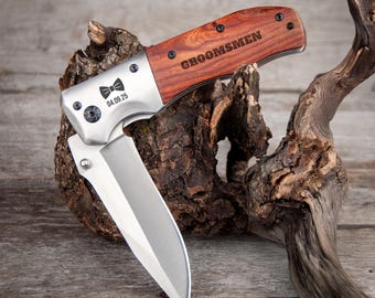 May include: A folding pocket knife with a wooden handle and a silver blade. The handle is engraved with the word "GROOMSMEN". The knife also features a bow tie graphic and the date "04.09.25". The knife is displayed on a piece of wood.