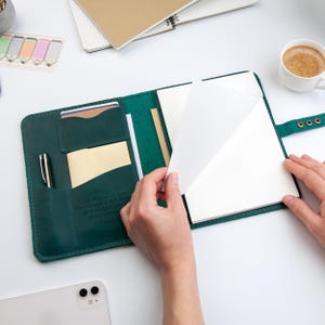 May include: A dark green leather notebook cover featuring multiple interior pockets for cards and documents, a pen holder, and a secure snap closure. The cover holds a stack of white paper sheets and includes an embossed inspirational quote.