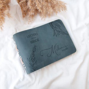 Pode incluir: Um di&aacute;rio de couro azul escuro com o texto "Cartas para a noiva" e um design floral. O di&aacute;rio &eacute; personalizado com o nome "Melanie" e a data "03.15.2024".