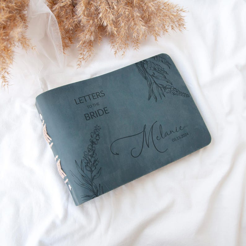 Pode incluir: Um di&aacute;rio de couro azul escuro com o texto "Cartas para a noiva" e um design floral. O di&aacute;rio &eacute; personalizado com o nome "Melanie" e a data "03.15.2024".